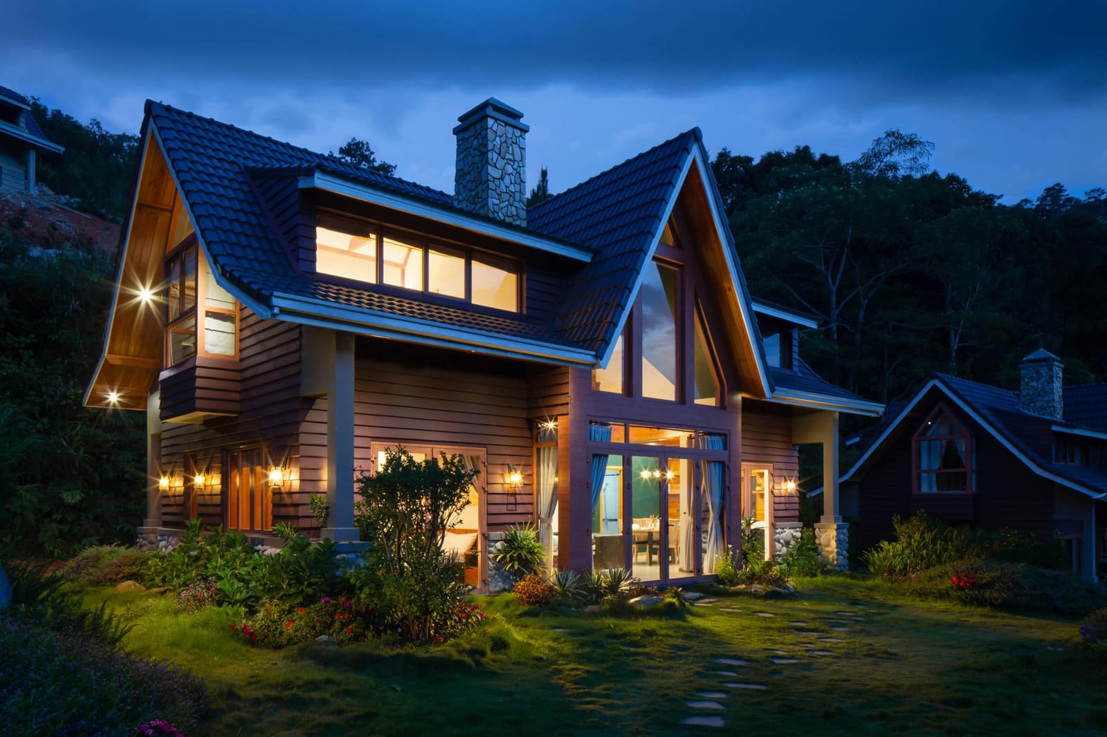 A two-story wooden house at dusk, with its windows lit and a garden in front